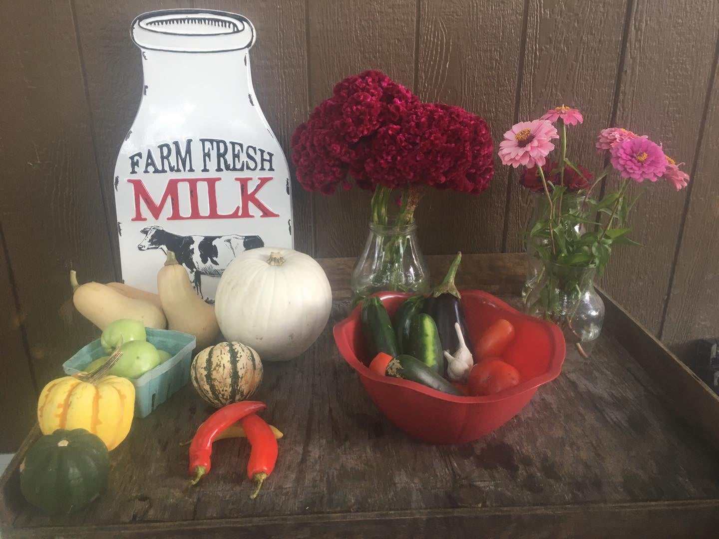 Farm fresh produce display featuring a variety of vegetables including squash, peppers, zucchini, and tomatoes, alongside a white pumpkin and green apples. Decorative flowers in glass vases are also present, with a vintage milk sign in the background, creating a rustic farm market ambiance.