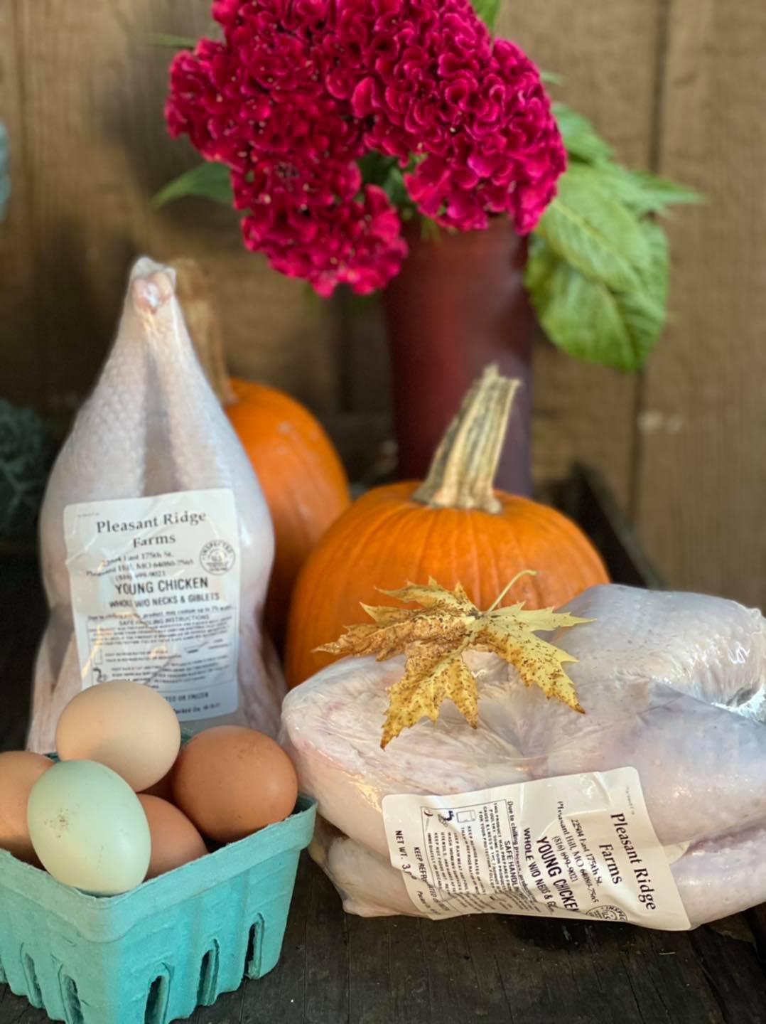Fresh farm produce featuring young chickens, eggs in a blue carton, vibrant pumpkins, and a bouquet of pink flowers, all set against a rustic wooden backdrop.
