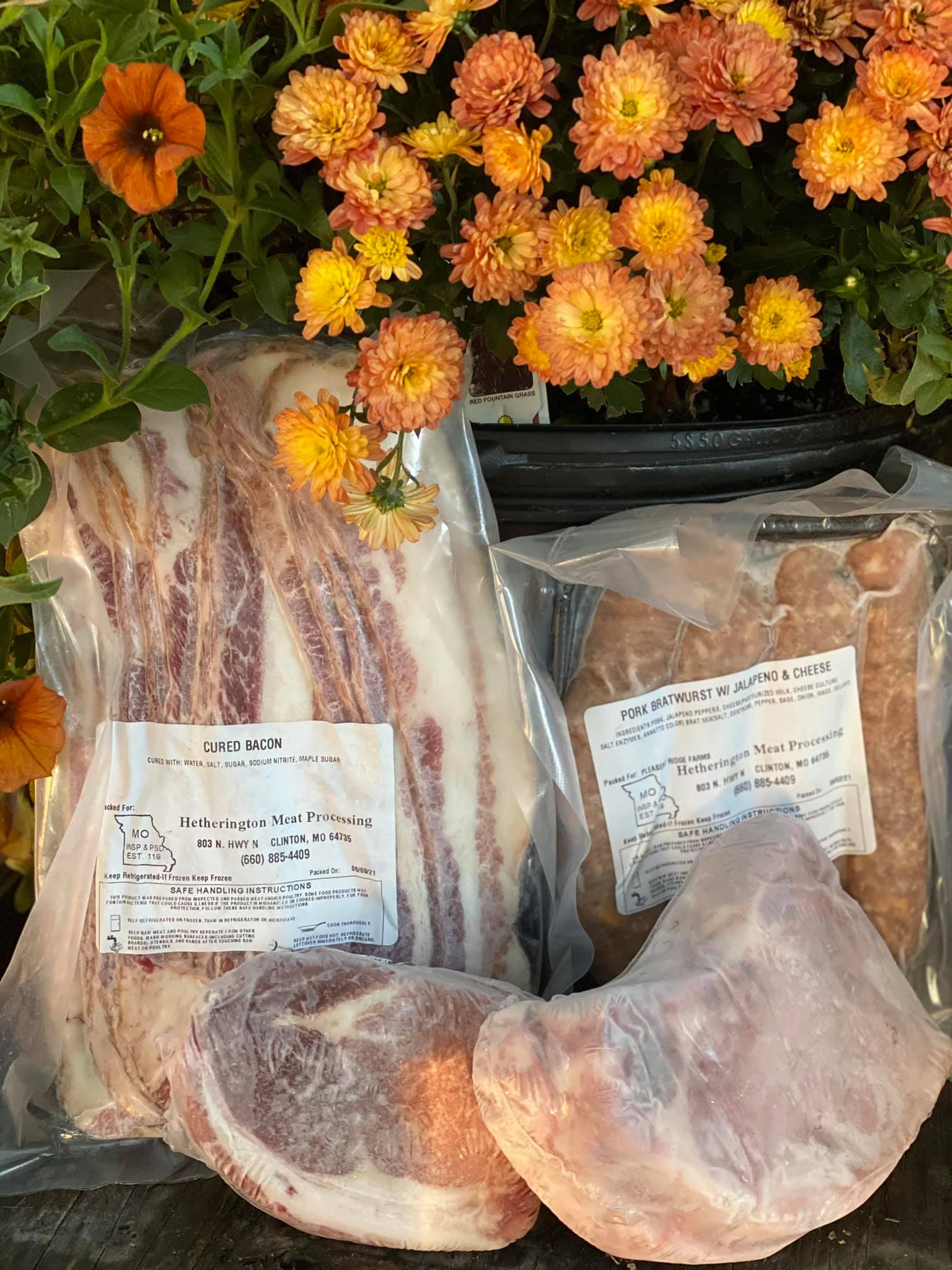 Assorted packaged meats from Hetherington Meat Processing, including cured bacon, pork bratwurst with jalapeño and cheese, and various cuts of pork, displayed alongside vibrant orange and yellow flowers, emphasizing local farm-fresh products.