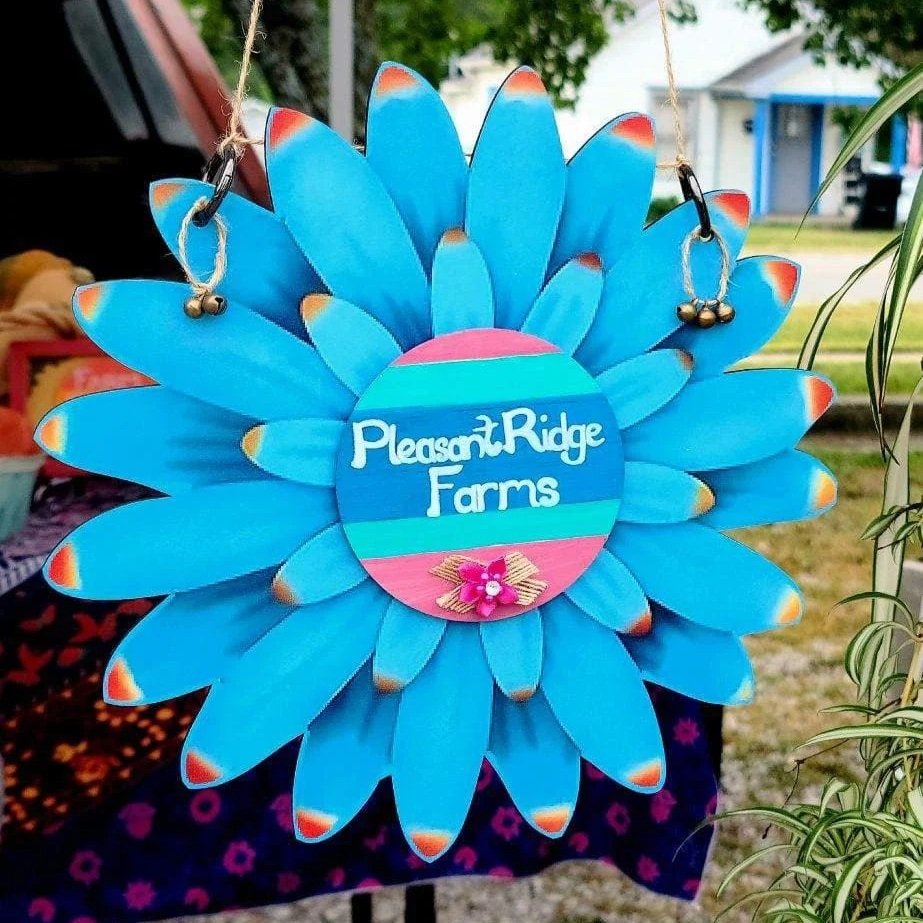 Colorful sign for Pleasant Ridge Farms featuring a large blue daisy design with a circular center displaying the farm's name, surrounded by decorative elements. Ideal for farm or market branding.