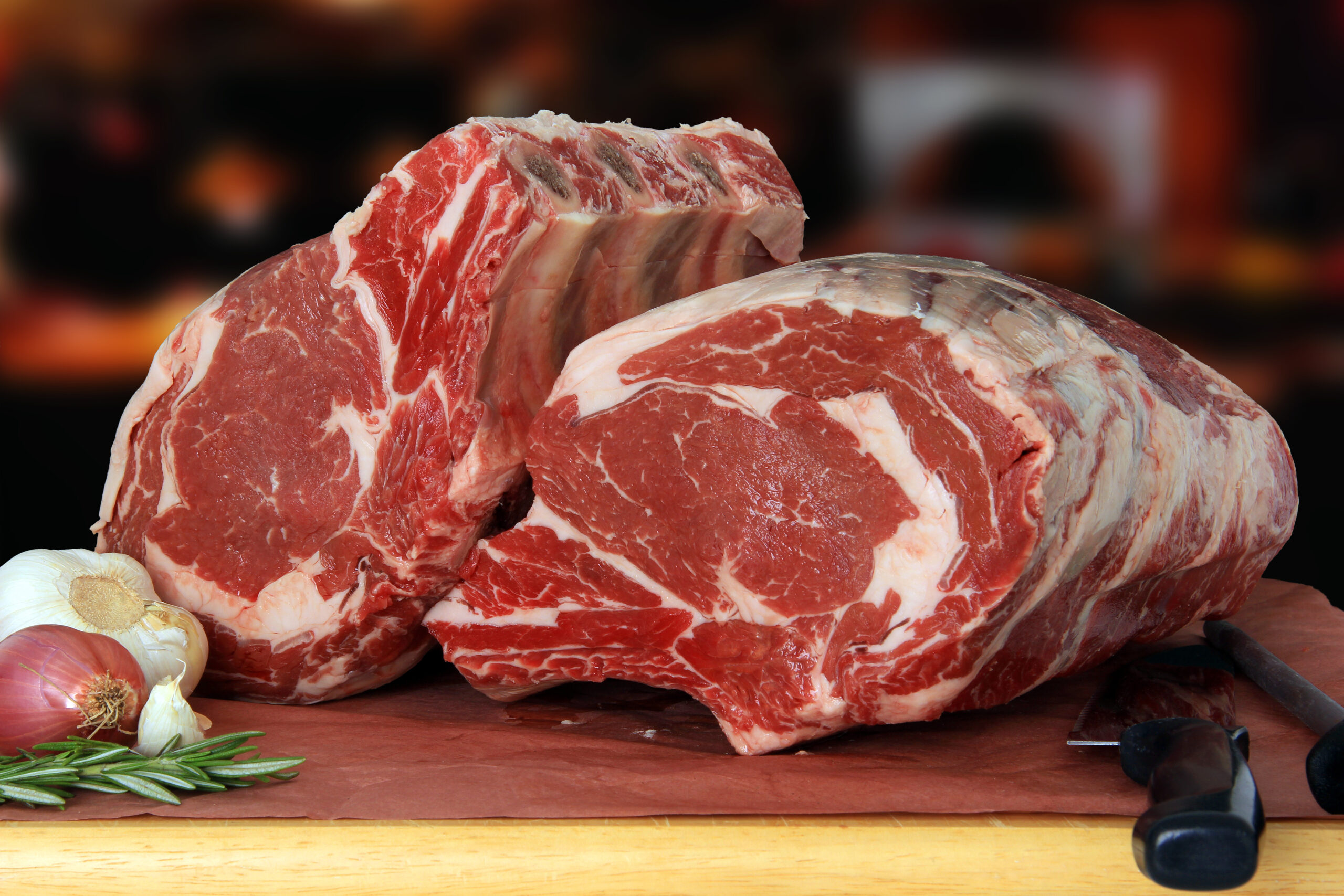 Fresh cuts of ribeye beef roast displayed on butcher paper, accompanied by garlic, shallots, and fresh rosemary, ideal for grilling or roasting.